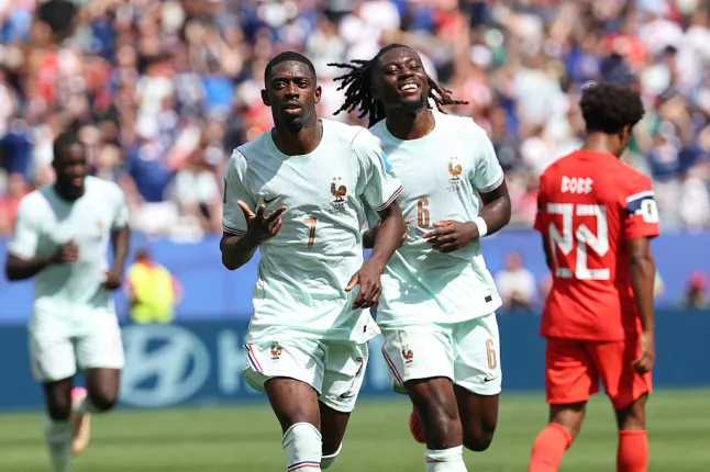 Two soccer players in light-blue jerseys celebrate a goal on a sunny stadium field, with fans in the stands behind them.