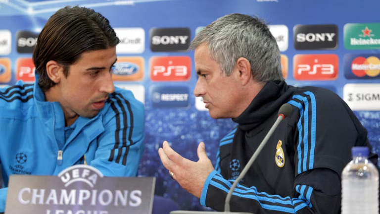Two men in Real Madrid gear speak at a UEFA Champions League press conference, facing each other and gesturing.