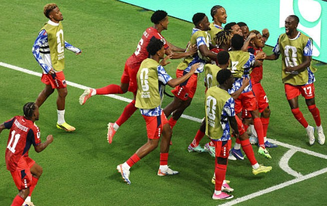 Soccer players in red uniforms celebrate on a green field, some wearing yellow bibs, as they hug and high-five near the penalty area.