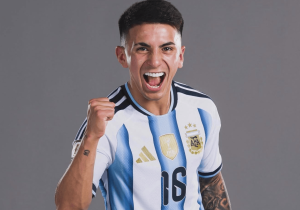 Argentinian soccer player in a light blue and white striped jersey celebrating with a raised clenched fist against a gray backdrop.
