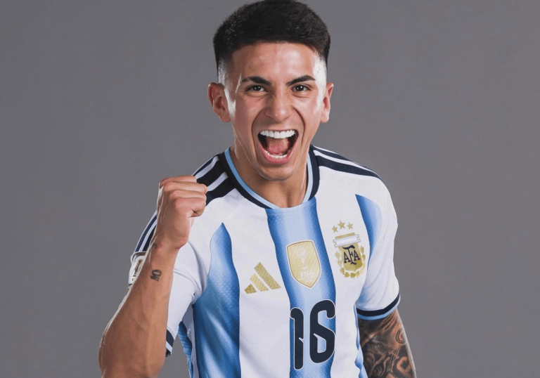 Argentinian soccer player in a light blue and white striped jersey celebrating with a raised clenched fist against a gray backdrop.