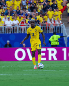 Soccer player in yellow jersey #23 controls ball on a green field with a crowded stadium in the background. Alternatives: A footballer wearing a yellow kit dribbles the ball during a match with fans in yellow in the stands.