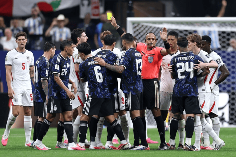 Soccer players from two teams surround the referee as he raises a yellow card during a stoppage on the field.