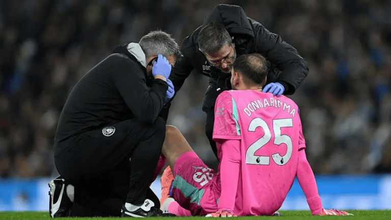 Medical staff attend to a pink-jersey goalkeeper sitting on the field after an injury, as coaches crouch nearby with concerned faces.