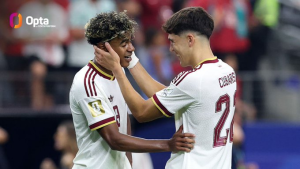 Two football players in white jerseys celebrate on the pitch, one cupping the other's face in a hug as teammates and fans blur in the background.