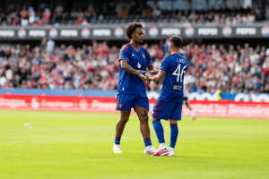 Two Manchester United players shake hands on a sunlit football pitch with a cheering crowd in the stands nearby.]','