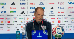 Man in England jersey at a press conference, speaking into a microphone with sponsor backdrop behind him; laptop shows a FIFA World Cup Miami graphic, with a soccer ball and water bottles on the table.