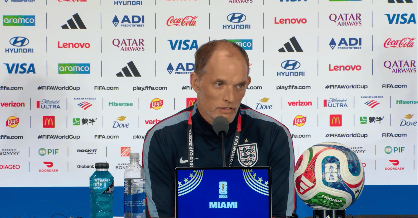 Man in England jersey at a press conference, speaking into a microphone with sponsor backdrop behind him; laptop shows a FIFA World Cup Miami graphic, with a soccer ball and water bottles on the table.