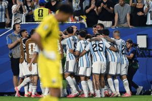 Argentine national team players in white-and-light-blue striped uniforms huddle on the field while cheering fans watch from the stands behind them