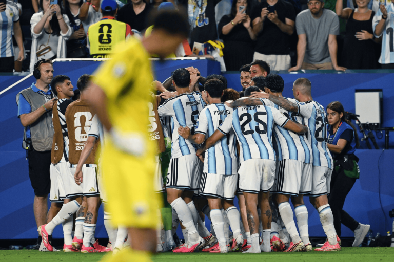 Argentine national team players in white-and-light-blue striped uniforms huddle on the field while cheering fans watch from the stands behind them