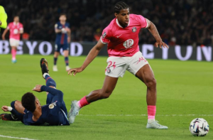 Soccer player in a pink kit dribbles the ball as a defender slides on the grass behind him.
