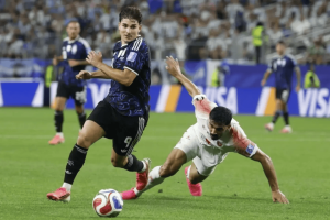 Soccer player in a dark blue jersey (number 9) controls the ball as a white-jersey opponent slides in for a tackle on the green field.