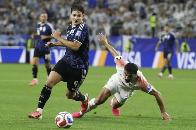 Soccer player in a dark blue jersey (number 9) controls the ball as a white-jersey opponent slides in for a tackle on the green field.