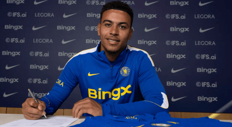 Young man in a Chelsea blue signing top signs a contract at a table during a signing event, backdrop covered with sponsor logos.