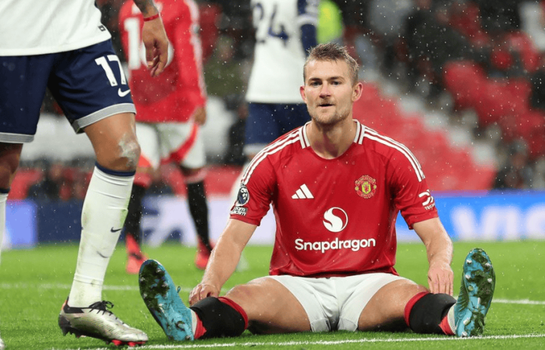 Manchester United player in red kit sits on the pitch after a challenge, with teammate legs nearby in white socks and cleats.
