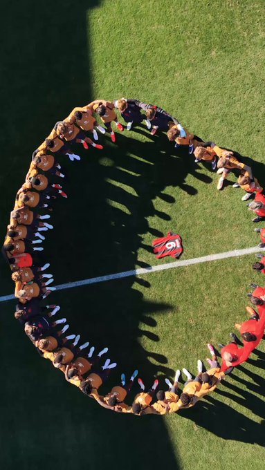 Overhead view of a diverse group standing in a circle on a grassy field, forming a ring around a red item on the ground, with long shadows cast by the sun.