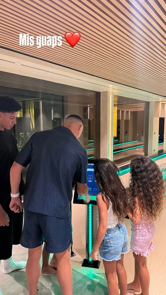 Man and two young girls interact with a blue touchscreen information kiosk in a bright, modern indoor hallway.