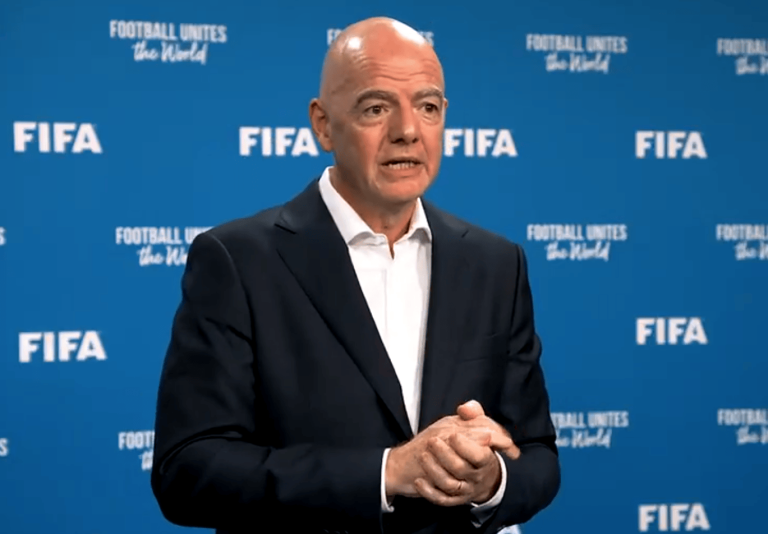 Bald man in a dark suit speaks at a FIFA press conference against a blue backdrop with FIFA logos.