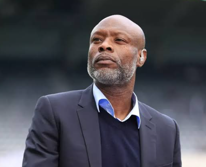 Bald man with a gray beard wearing a dark suit and blue shirt, looking upward with a serious expression at an outdoor venue