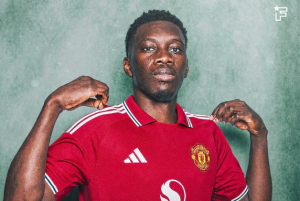 Portrait of a man in a red Manchester United jersey posing with hands at his shoulders against a green textured background.
