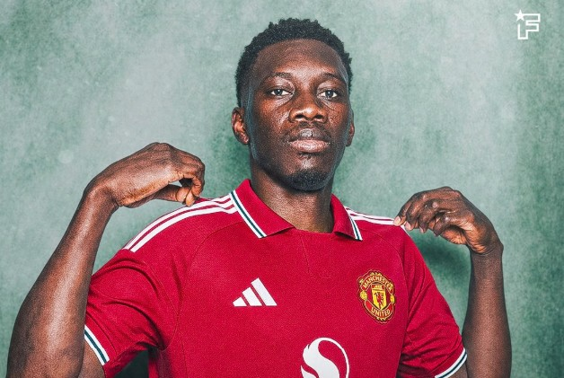 Portrait of a man in a red Manchester United jersey posing with hands at his shoulders against a green textured background.