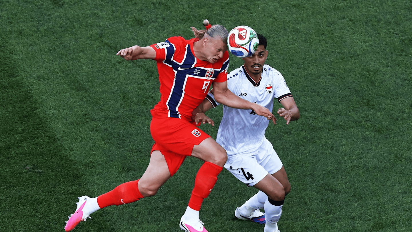 Two soccer players clash for the ball in midair on a green pitch, one in red and the other in white.