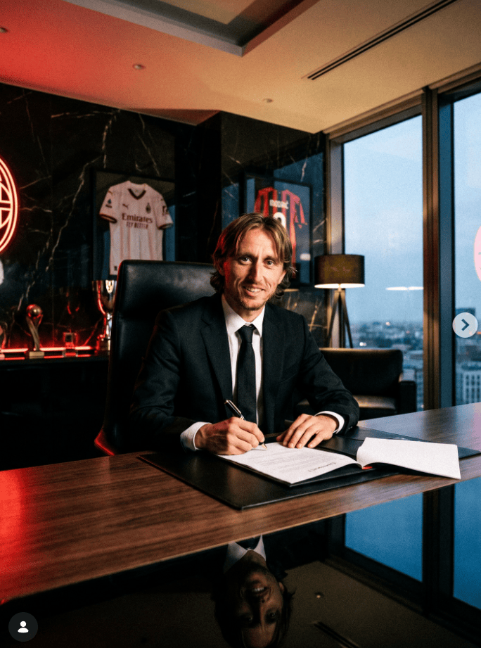 Businessman in a black suit signs documents at a wooden desk in a modern office, with framed sports jerseys in the background.