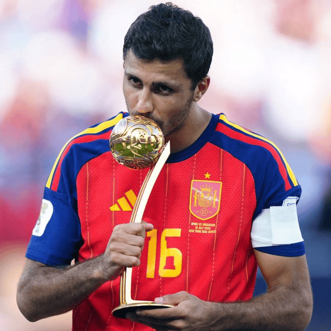 Soccer player in a red Spain jersey kissing the Ballon d'Or trophy on a field side view, wearing captain armband on his left arm