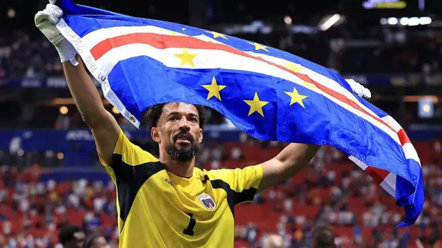Goalkeeper in a yellow jersey proudly holds up a blue flag with yellow stars in a stadium, celebrating.