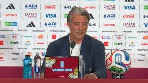 Man in a navy blazer speaks into a microphone at a press conference with a FIFA backdrop and 'Vancouver' on a tablet in front of him, soccer ball on the right side.