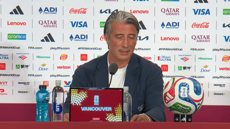 Man in a navy blazer speaks into a microphone at a press conference with a FIFA backdrop and 'Vancouver' on a tablet in front of him, soccer ball on the right side.