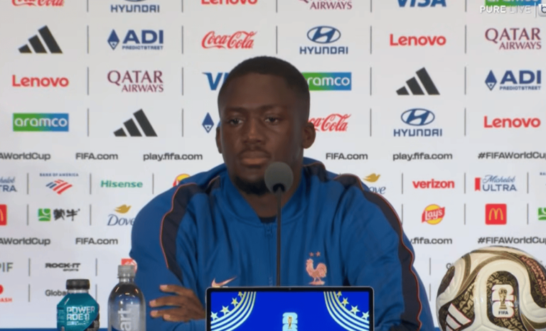 Male footballer in a blue jacket sits at a press conference table with a microphone, backdrop covered in sponsor logos and a FIFA World Cup ball to the right.