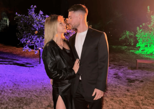 A couple shares a kiss outdoors at night, standing close under colorful purple and green lights.