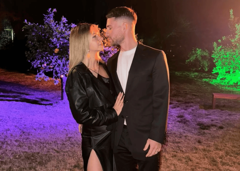 A couple shares a kiss outdoors at night, standing close under colorful purple and green lights.