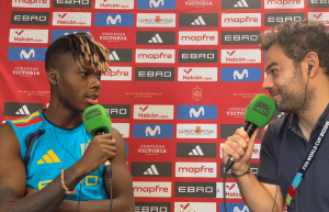 Two men interview on a red sponsor backdrop; left is a Black football player in a blue jersey with braided hair, speaking into a green microphone, while the interviewer on the right listens, holding a green mic.