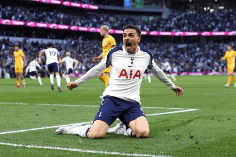 Soccer player in a white Tottenham jersey slides on knees, arms spread, celebrating a goal on the pitch.