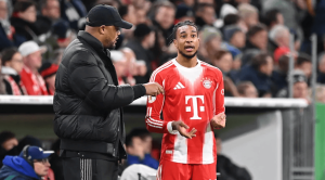 Soccer coach in a black puffer jacket talks to a Bayern Munich player in a red and white jersey on the sideline during a match.