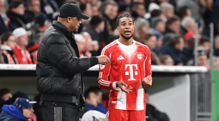 Soccer coach in a black puffer jacket talks to a Bayern Munich player in a red and white jersey on the sideline during a match.