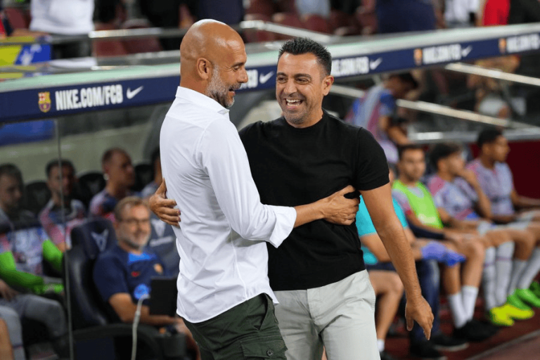 Two men share a hug and smile on the soccer sideline, with players seated in the dugout behind them.