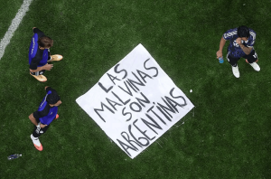 Aerial view of three soccer players around a white banner on green grass that reads 'Las Malvinas son Argentinas'.