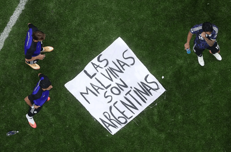 Aerial view of three soccer players around a white banner on green grass that reads 'Las Malvinas son Argentinas'.