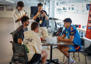Group interview in a bright open space: a footballer in a blue kit sits at a table answering questions while teammates and crew with cameras and phones stand and listen nearby.