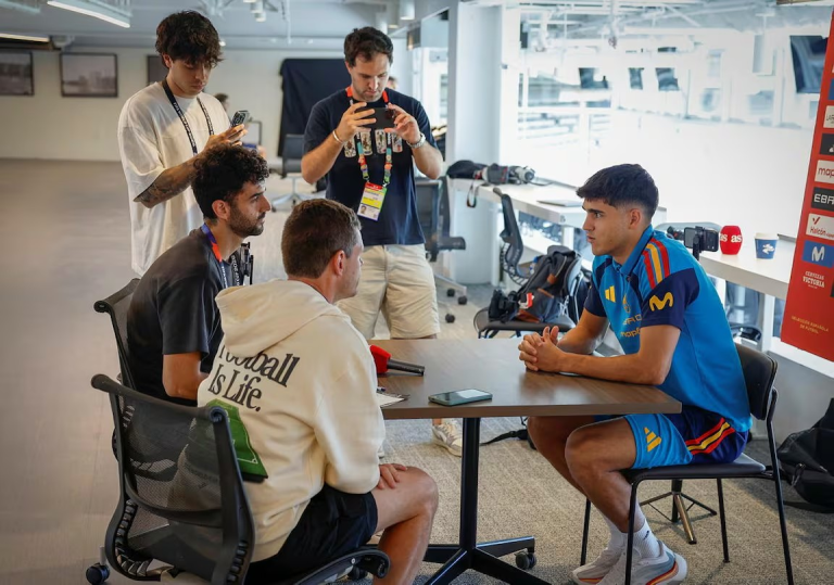 Group interview in a bright open space: a footballer in a blue kit sits at a table answering questions while teammates and crew with cameras and phones stand and listen nearby.