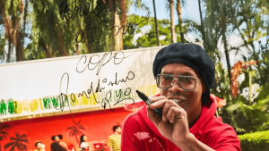Man with large glasses and a black beret writes on a transparent board outdoors, marker in hand, colorful background nearby.