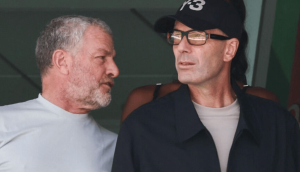 Two men engaged in conversation: one with a gray beard in a white shirt, the other wearing a cap, glasses, and a dark jacket.