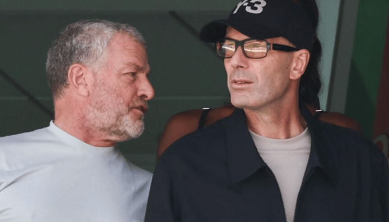 Two men engaged in conversation: one with a gray beard in a white shirt, the other wearing a cap, glasses, and a dark jacket.