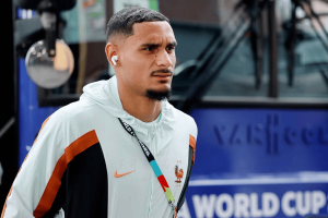 Portrait of a man wearing a light jacket with orange and black color blocks, white AirPods, and a multicolor lanyard. He stands near a blue bus, looking off to the side.