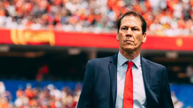 Soccer manager in a navy suit and red tie standing on the sideline with a stadium crowd in the background, focused on the match.