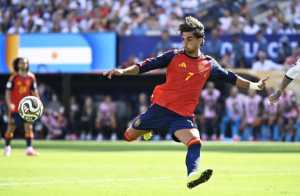 Soccer player in a red Spain jersey mid-kick during a match, ball near his foot on a green field with a crowd in the background.