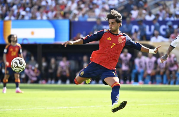 Soccer player in a red Spain jersey mid-kick during a match, ball near his foot on a green field with a crowd in the background.